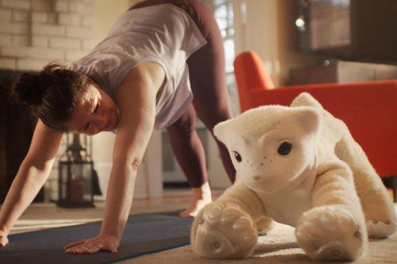 A woman in a white sleeveless top and maroon leggings is doing a downward dog yoga pose on a mat indoors. Beside her there is a large, cream-colored robotic creature with black eyes and a soft, plush-like texture, positioned similarly on the floor. The background includes a red chair and a softly lit room.