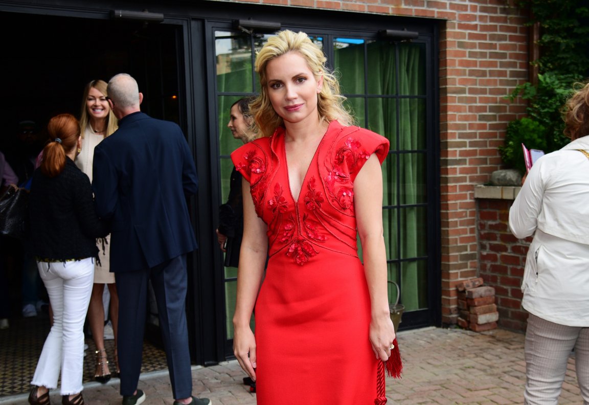 A woman with blonde hair is wearing a red dress with intricate floral embroidery and a deep V-neckline. She is standing outside in front of a building with brick walls and large black-framed glass doors with green curtains behind them. Several other people are visible in the background, engaged in conversation.