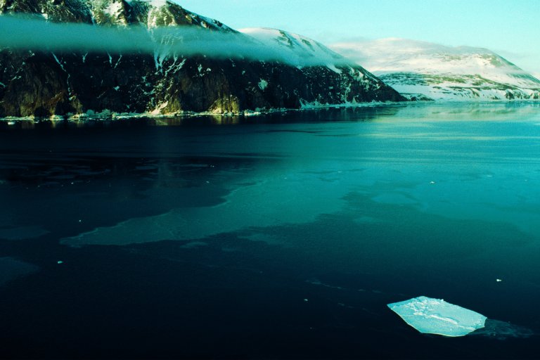 Snow-covered mountains with rocky faces are reflected in a calm, icy blue body of water, with a small floating ice chunk visible in the foreground. A thin layer of mist or low clouds stretches horizontally across the middle of the mountains.