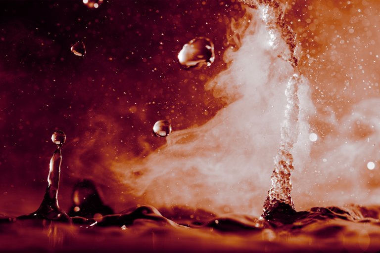 Close-up of water droplets splashing and rising with mist or steam in a warm, reddish-orange light. The image captures the dynamic motion and texture of the liquid and vapor.