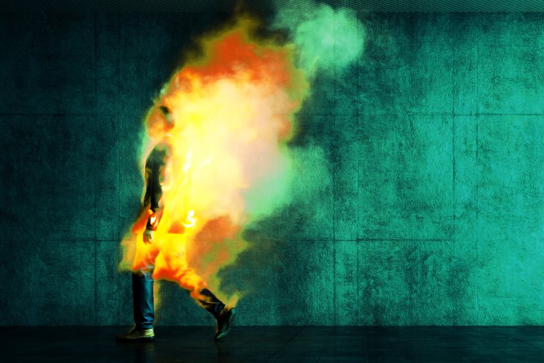 A person walking with their body engulfed in bright flames and smoke, creating a striking contrast against a dark teal textured wall and floor background. The fire emits intense yellow, orange, and red hues, blending into greenish smoke. The person's silhouette is partially visible through the flames.