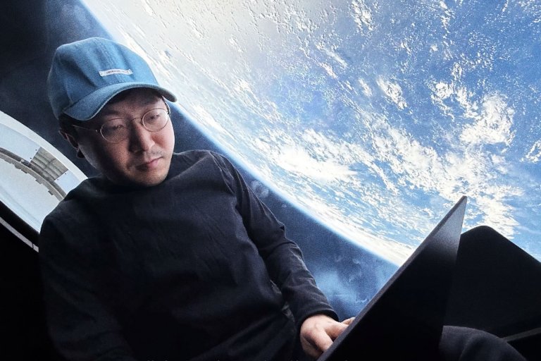 A man wearing glasses, a light blue cap, and a black long-sleeve shirt is sitting and using a laptop with the Earth visible through a large window behind him, suggesting he is in a spacecraft or space station.