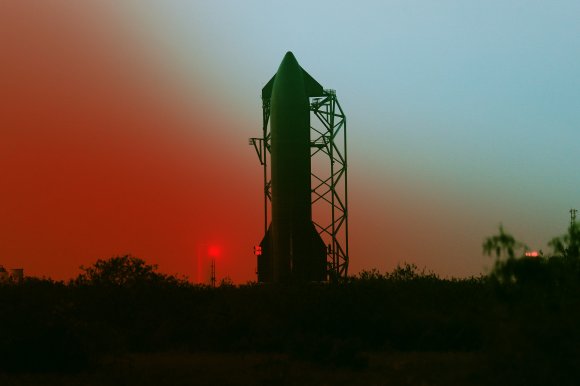 A large rocket stands vertically on a launch pad structure during twilight, with a gradient sky transitioning from red near the horizon to blue higher up. The silhouette of trees and bushes is visible in the foreground.
