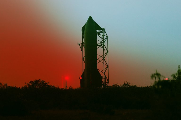 A large rocket stands vertically on a launch pad structure during twilight, with a gradient sky transitioning from red near the horizon to blue higher up. The silhouette of trees and bushes is visible in the foreground.
