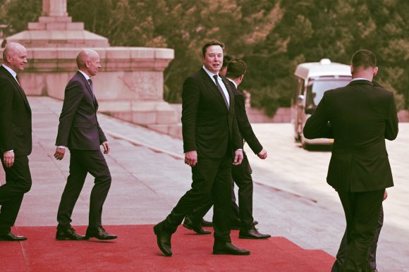 Elon Musk walking on a red carpet outdoors, accompanied by several men in black suits. The background includes stone steps and greenery, with a white vehicle visible in the distance.