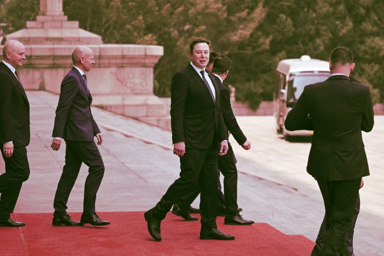 Elon Musk walking on a red carpet outdoors, accompanied by several men in black suits. The background includes stone steps and greenery, with a white vehicle visible in the distance.