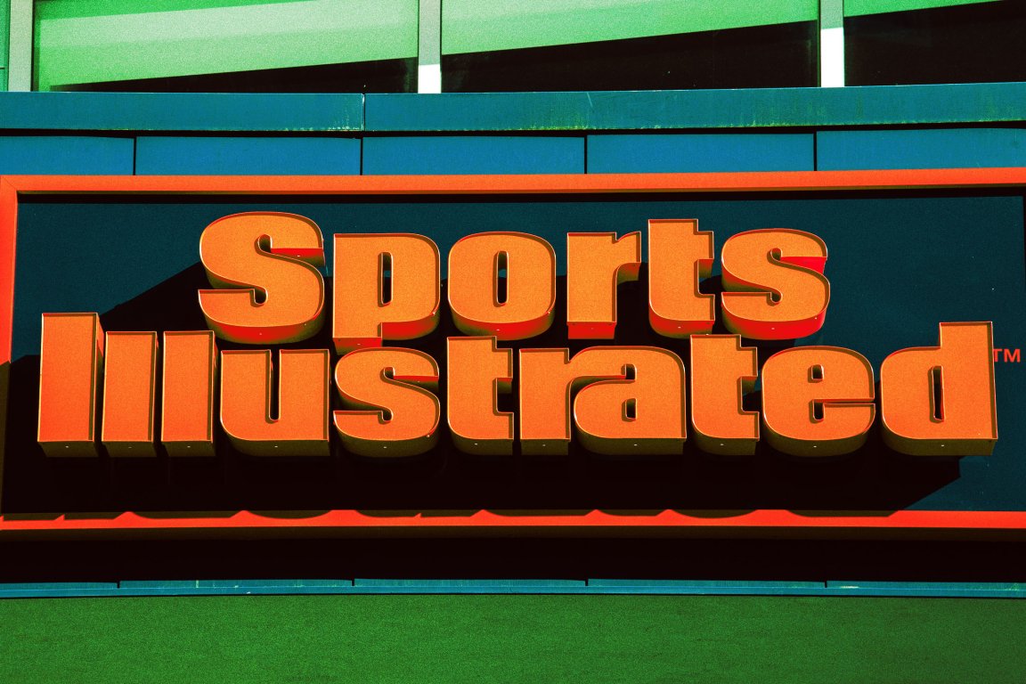 Large, bold, orange 3D letters spelling "Sports Illustrated" on a dark background with an orange border, mounted on a building facade with green and blue panels above and below.