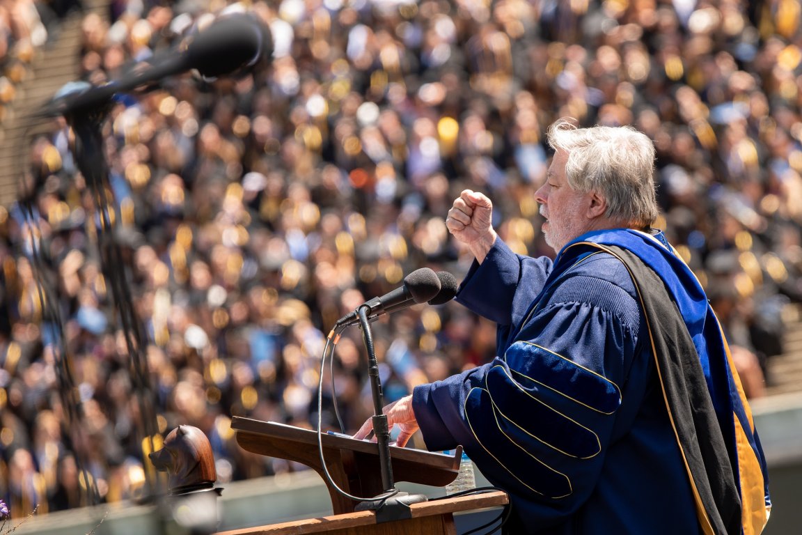 Steve Wozniak is speaking passionately at a podium, wearing a blue academic gown with black and gold trim. He is gesturing with his right hand raised in a fist. In front of him are two microphones, and a large crowd of people dressed in academic regalia is visible in the blurred background.