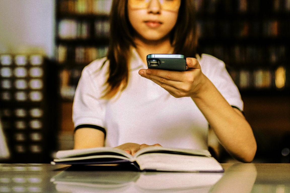 A person wearing a white shirt with black trim is sitting at a table with an open book. They are holding a smartphone in one hand, positioned above the book, and their face is partially visible but out of focus. The background appears to be a library or a room with bookshelves.