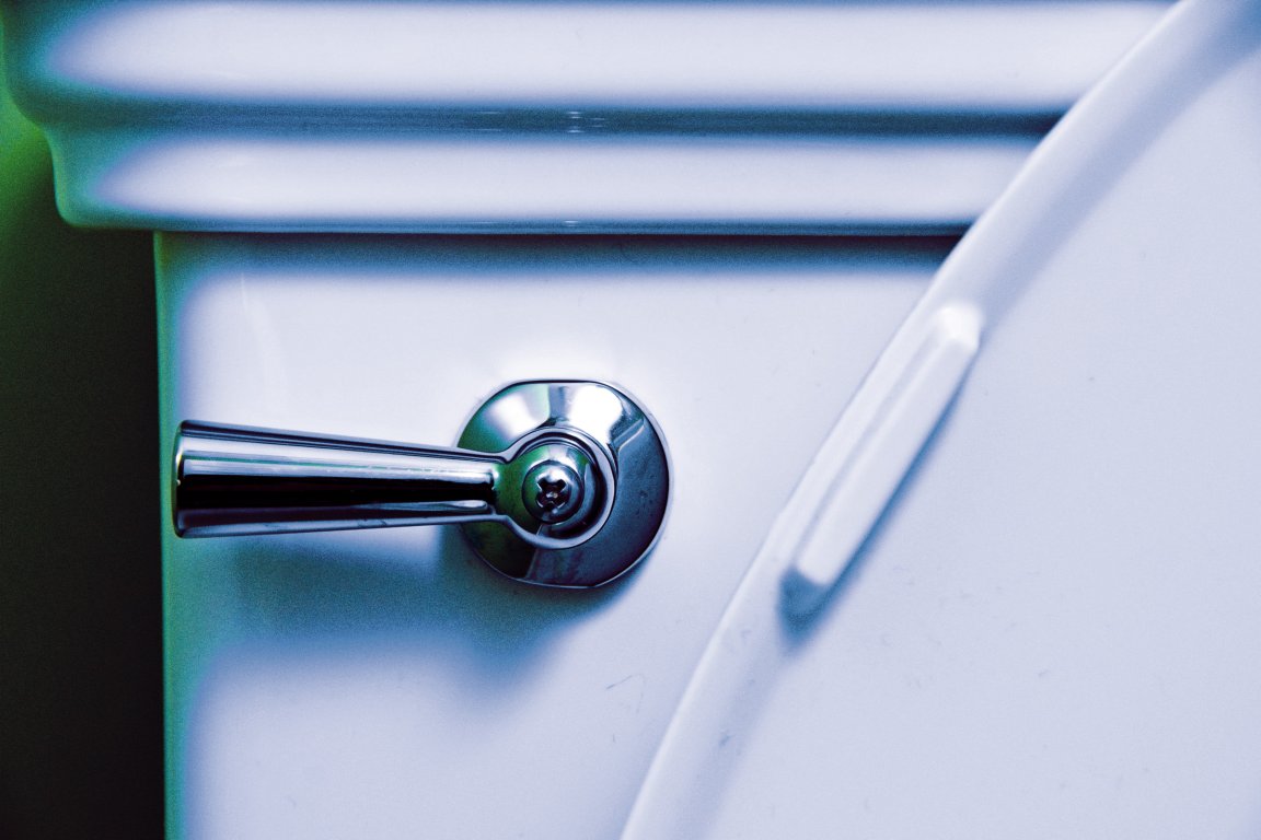 Close-up of a white toilet tank with a shiny chrome flush handle on the left side and a partially visible toilet seat on the right.