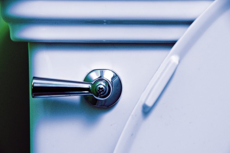 Close-up of a white toilet tank with a shiny chrome flush handle on the left side and a partially visible toilet seat on the right.
