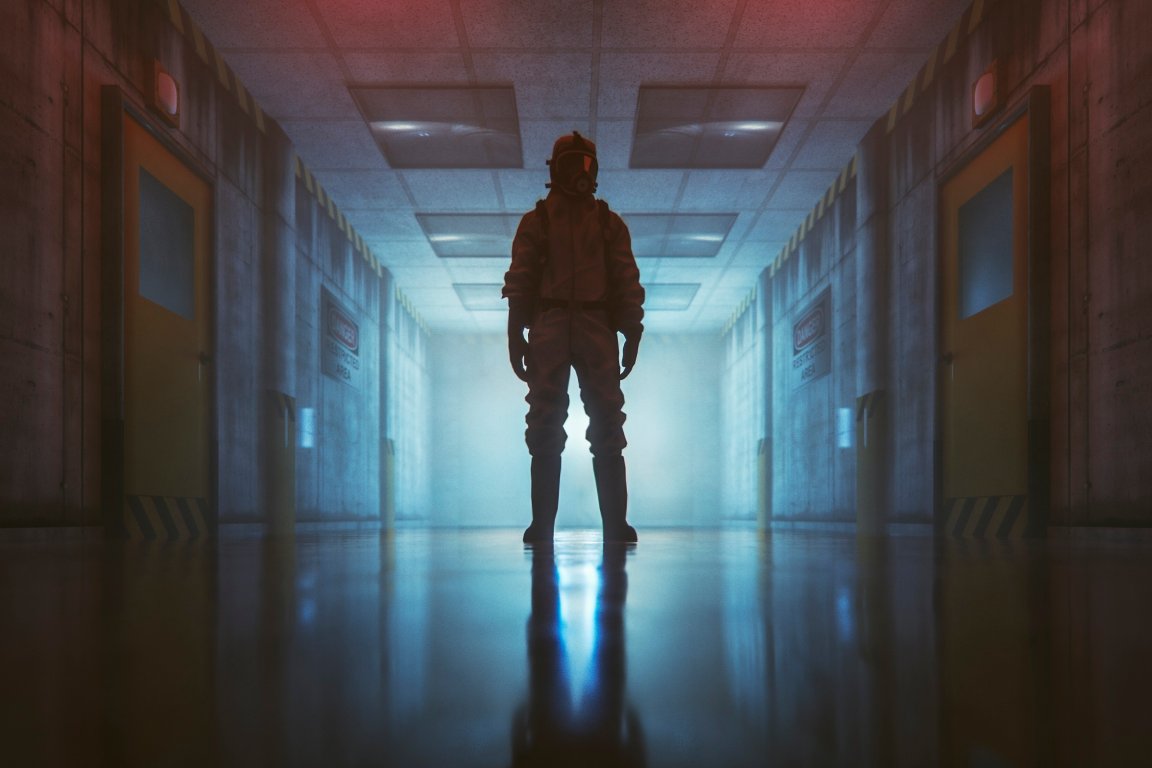 A person wearing a full protective hazmat suit and gas mask stands in the middle of a dimly lit, industrial hallway.