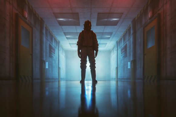 A person wearing a full protective hazmat suit and gas mask stands in the middle of a dimly lit, industrial hallway.
