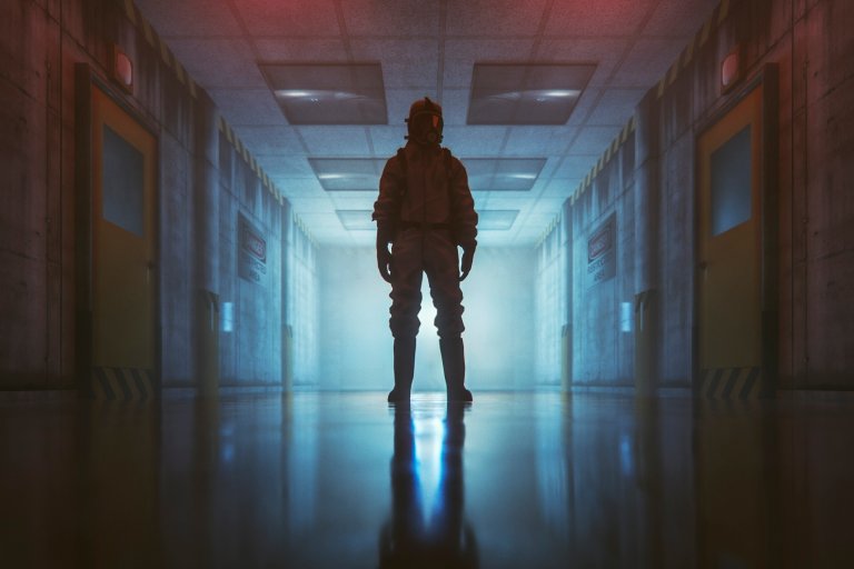 A person wearing a full protective hazmat suit and gas mask stands in the middle of a dimly lit, industrial hallway.
