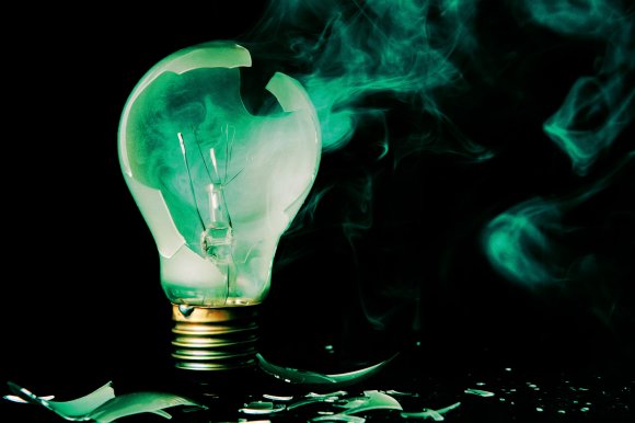 A broken light bulb with shattered glass pieces scattered around its base, emitting greenish smoke against a black background.