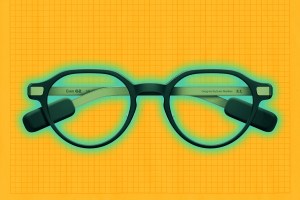 Round black Even Realities G2 live-caption eyeglasses with thick frames and attached black rectangular sensors on the temples, placed on an orange grid background.
