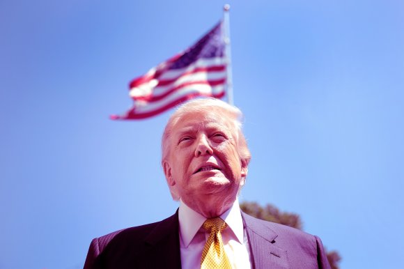 A close-up of a Donald Trump wearing a dark suit, white shirt, and yellow patterned tie, standing outdoors with a clear blue sky and a waving American flag in the background.