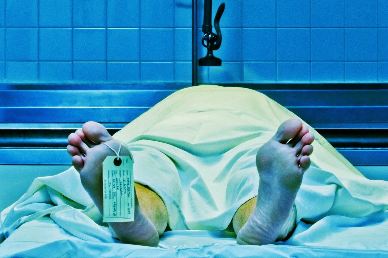 A body covered with a white sheet lies on a metal table in a morgue, with a toe tag attached to the left foot. The background features blue-tiled walls and a metal fixture above the body.