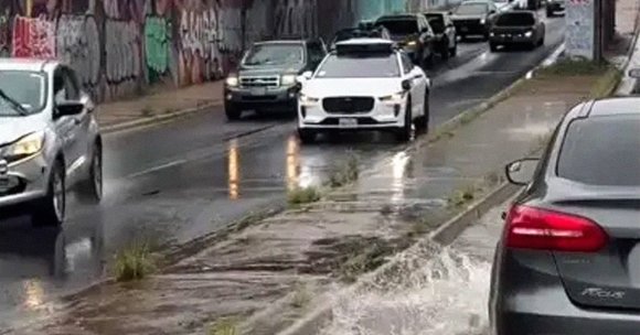A wet urban street with several cars driving and parked along the road. The road surface is wet, reflecting the headlights of the vehicles, and there is water splashing near the curb, indicating recent rain or flooding. A Waymo self-driving taxi is in the roadway.