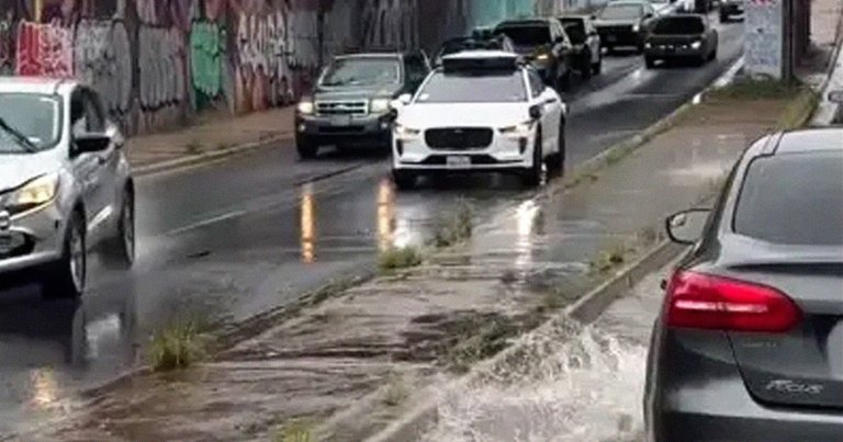 A wet urban street with several cars driving and parked along the road. The road surface is wet, reflecting the headlights of the vehicles, and there is water splashing near the curb, indicating recent rain or flooding. A Waymo self-driving taxi is in the roadway.