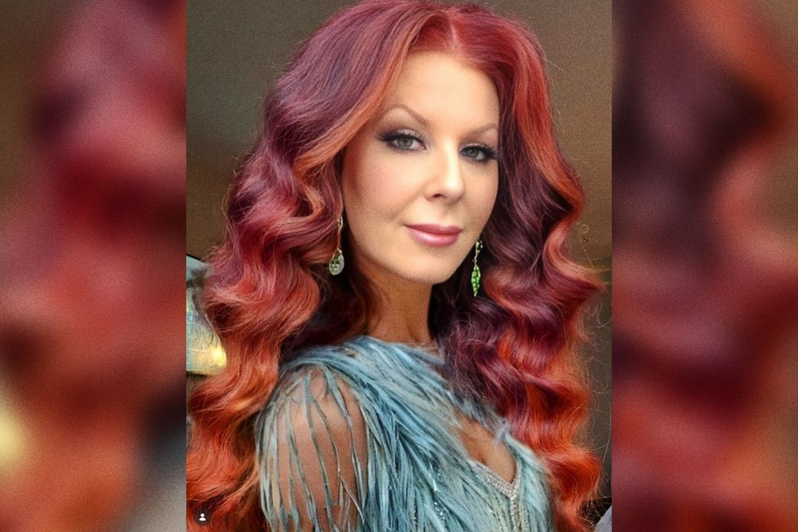 Woman with long, wavy red hair wearing a light blue, fringed top and dangling earrings, looking directly at the camera with a slight smile.