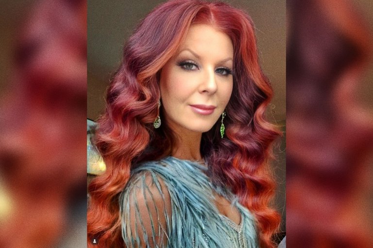 Woman with long, wavy red hair wearing a light blue, fringed top and dangling earrings, looking directly at the camera with a slight smile.
