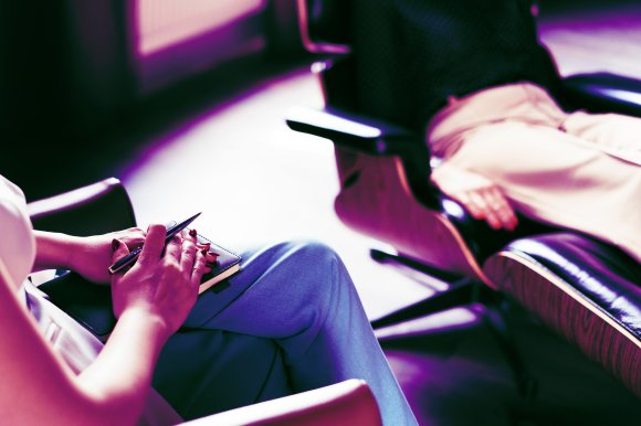 Close-up of two people sitting in modern chairs, one person holding a pen and notebook on their lap, the other person reclining with hands resting on their lap.