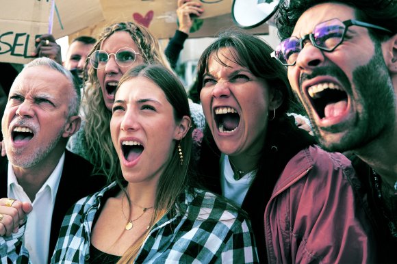 A diverse group of five people shouting passionately, likely at a protest or rally. They appear intense and determined, with some holding signs in the background. The group includes men and women of different ages, with one person wearing glasses and another wearing a checkered shirt. The expressions convey strong emotion and unity.