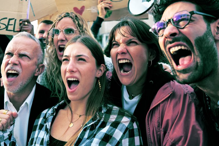 A diverse group of five people shouting passionately, likely at a protest or rally. They appear intense and determined, with some holding signs in the background. The group includes men and women of different ages, with one person wearing glasses and another wearing a checkered shirt. The expressions convey strong emotion and unity.