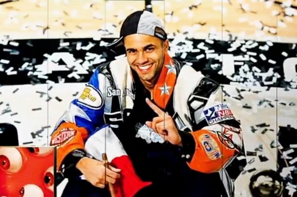 A man wearing a colorful jacket with various patches, including one that says "NBA CHAMPIONS," and a two-tone cap worn backward is smiling and holding up one finger. Confetti is falling around him, suggesting a celebratory moment.