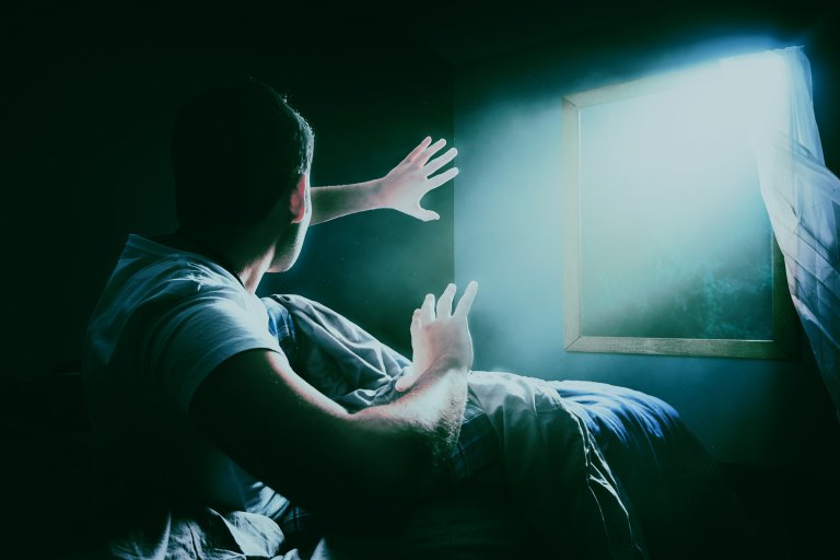 A photo illustration featuring a man in bed frightened by a light shining through his window from outside.