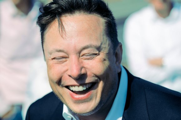 Close-up of a man with short dark hair laughing with his eyes closed, wearing a dark jacket and a white shirt. The background is blurred with indistinct figures.