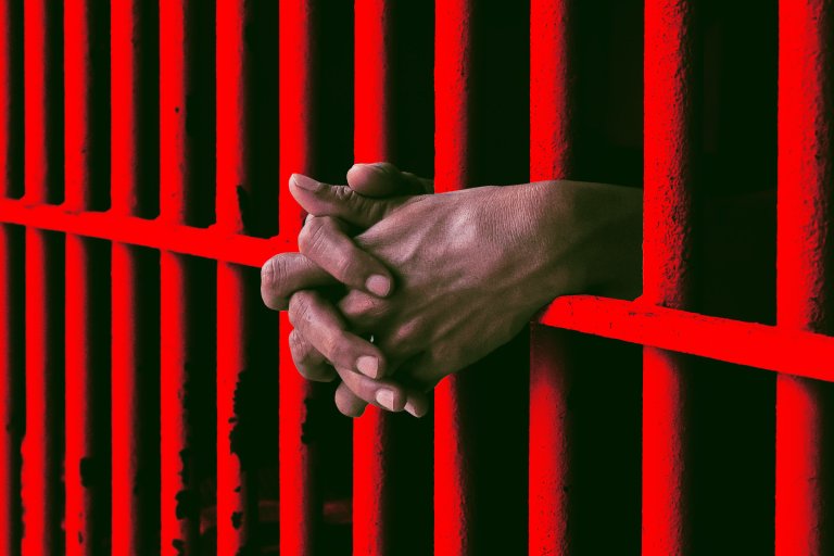 A color-treated photo illustration featuring a prisoner's hands folded through the bars of his cell, bathed in red.