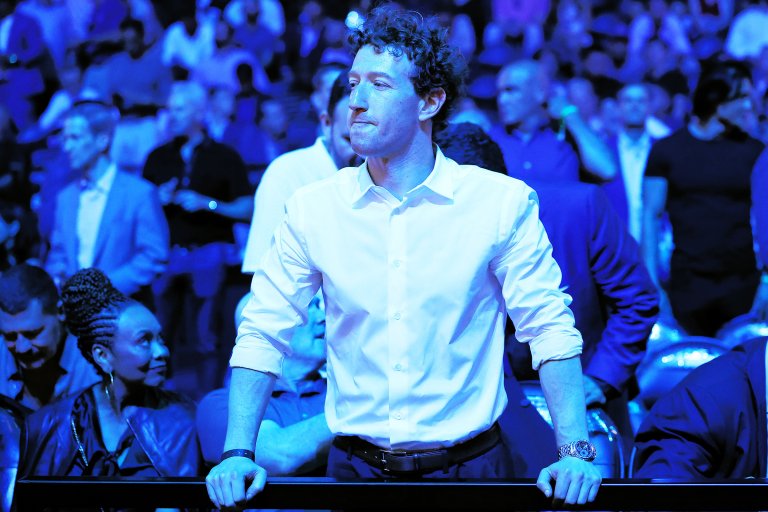 A man with curly hair wearing a white button-up shirt with rolled-up sleeves stands with his hands resting on a railing. He is looking to his left with a thoughtful expression. The background is filled with a crowd of people, all bathed in blue lighting.