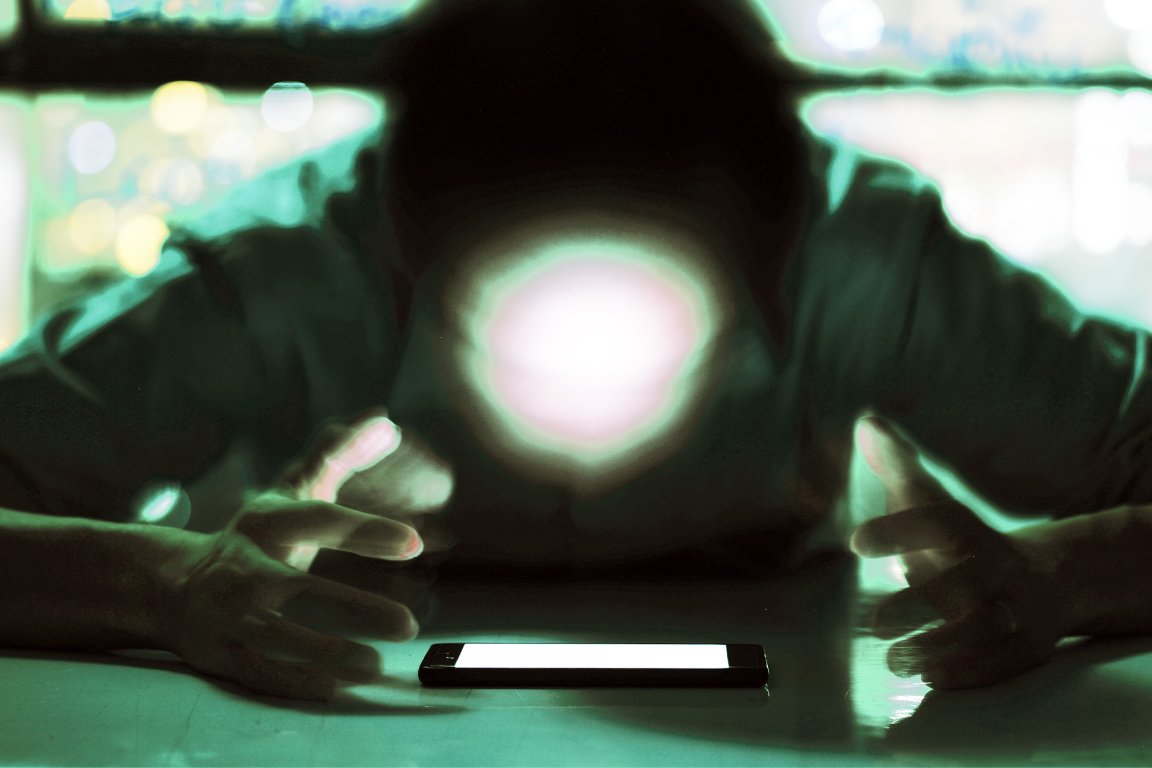 A color-treated lone exposure photograph of a person hovering over their phone screen, their illuminated face rendered as a featureless blur.