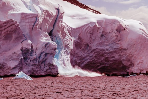 A large glacier with rugged, textured ice formations in shades of white and light blue, meeting a body of water. The water has a reddish tint, and there is mist or spray where the glacier touches the water. The sky above is partly cloudy.