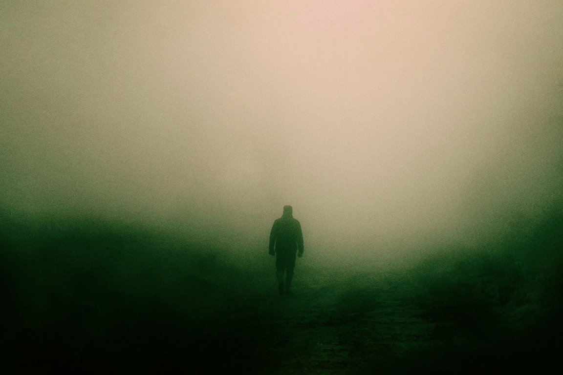 A lone figure walks through thick fog in the countryside.