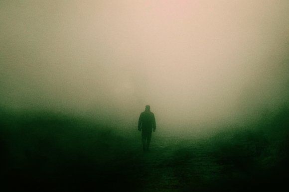 A lone figure walks through thick fog in the countryside.
