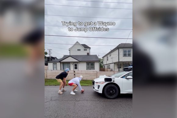 A screenshot featuring two young men in football uniforms attempting to play football with a Waymo.