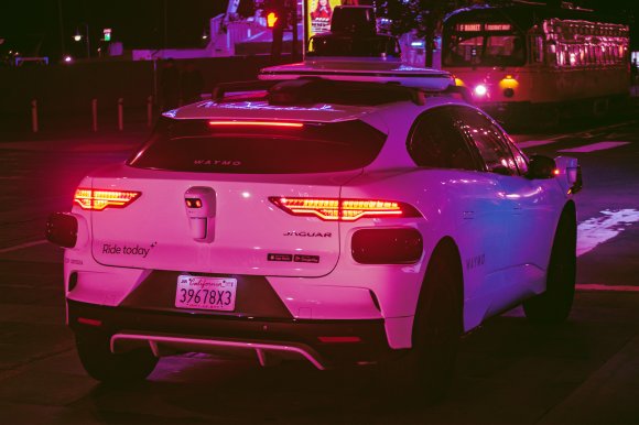 White Jaguar electric vehicle equipped with Waymo autonomous driving sensors on the roof and rear, parked on a city street at night with illuminated taillights and a California license plate. The car has "Ride today" and app store stickers on the back. A tram is visible in the background.