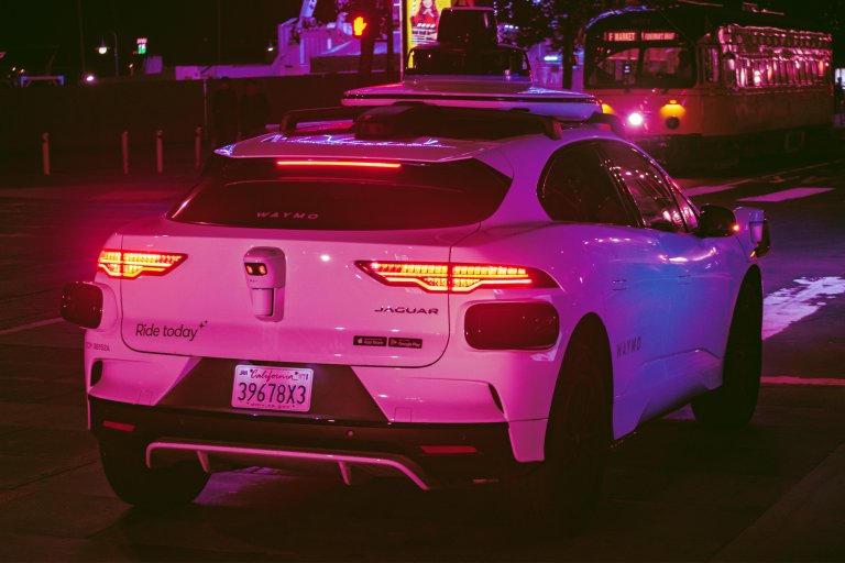 White Jaguar electric vehicle equipped with Waymo autonomous driving sensors on the roof and rear, parked on a city street at night with illuminated taillights and a California license plate. The car has "Ride today" and app store stickers on the back. A tram is visible in the background.