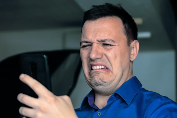 A man wearing a blue shirt is looking at a smartphone with a disgusted and grimacing facial expression.