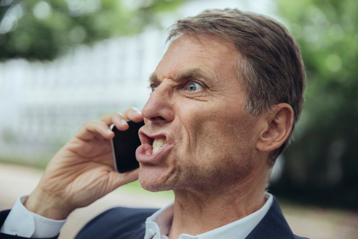 A middle-aged man with short brown hair is holding a smartphone to his ear and appears to be angrily shouting or arguing.