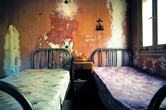 A photograph of a rundown, gross hotel that appears abandoned with two twin beds stripped of their sheets and a telephone between them.