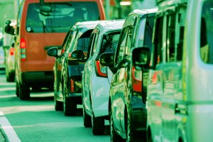 A line of cars and vans closely packed together on a road, viewed from the rear side. The vehicles include a white car, a black car, and an orange van, among others, reflecting light and nearby surroundings. The scene suggests traffic congestion.