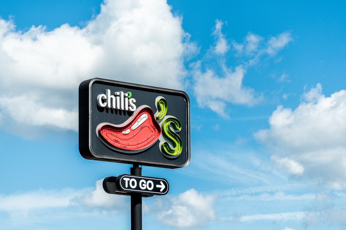 Chili's restaurant sign featuring a red chili pepper and the text "Chili's" on a black background, with an additional "TO GO" sign below it, set against a blue sky with scattered white clouds.