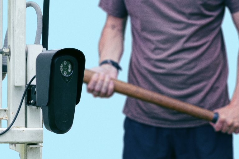 A composite photo illustration comically showing a Flock camera in the foreground and a man holding a baseball back in the background.