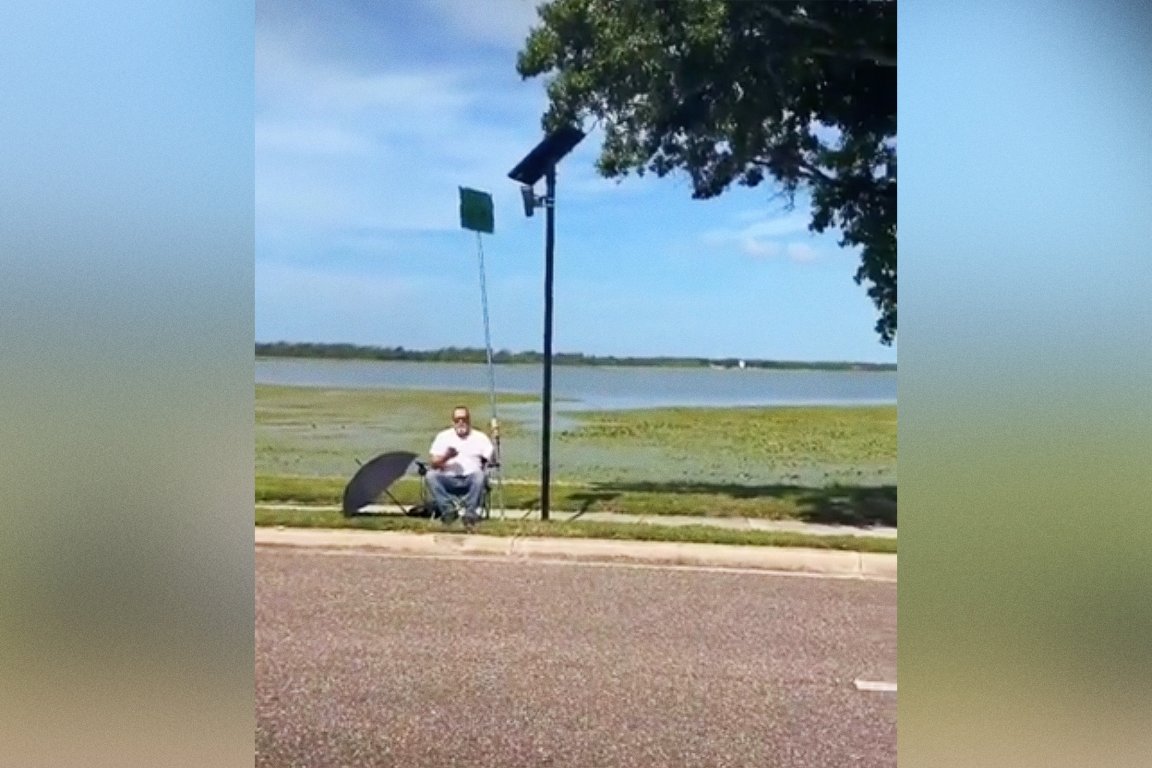 A screenshot of a video featuring a man sitting in a foldout chair next to a Flock camera on the side of a rural road, holding a single sign on a long pole that blocks the camera's view.
