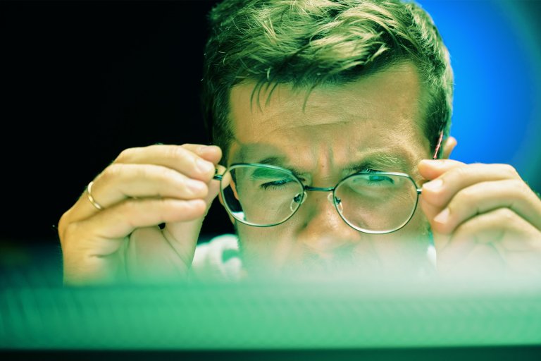 A color-treated image of a man correcting his glasses as he looks disbelievingly at his computer monitor.