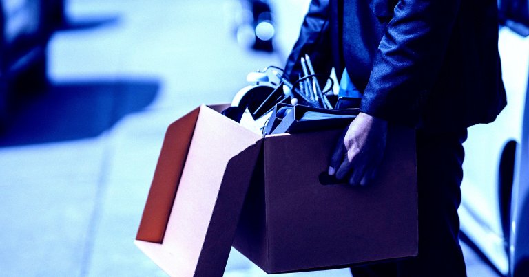 A person wearing a dark suit is holding an open cardboard box filled with office items, including a clock, pens in a holder, and a binder. The background is blurred, suggesting an outdoor urban setting.